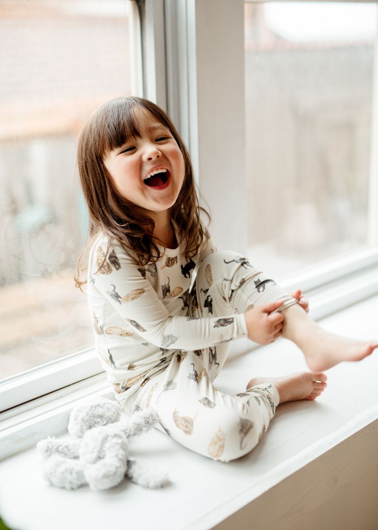 Little girl laughing and adjusting her pant leg while wearing Milkbarn’s Cat print cotton-spandex long sleeve two-piece pajama set.