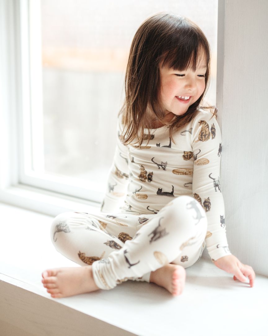 Little girl with dark hair smiling and looking to the side while sitting on a windowsill wearing Milkbarn’s cotton-spandex Cat print long sleeve two-piece pajama set.