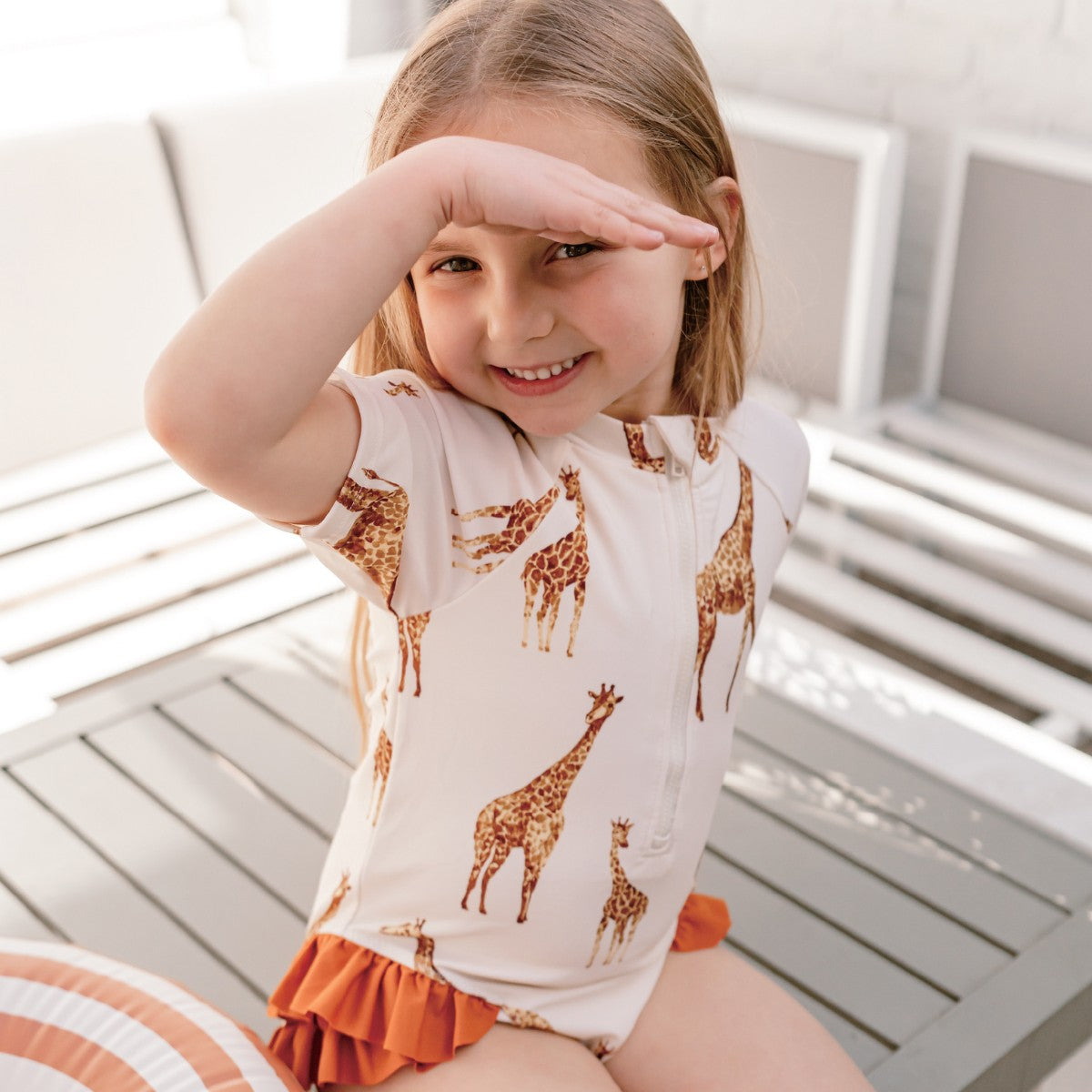 Toddler girl sitting by the pool edge wearing the Giraffe Zipper Short Sleeve Ruffle Leg Swimsuit