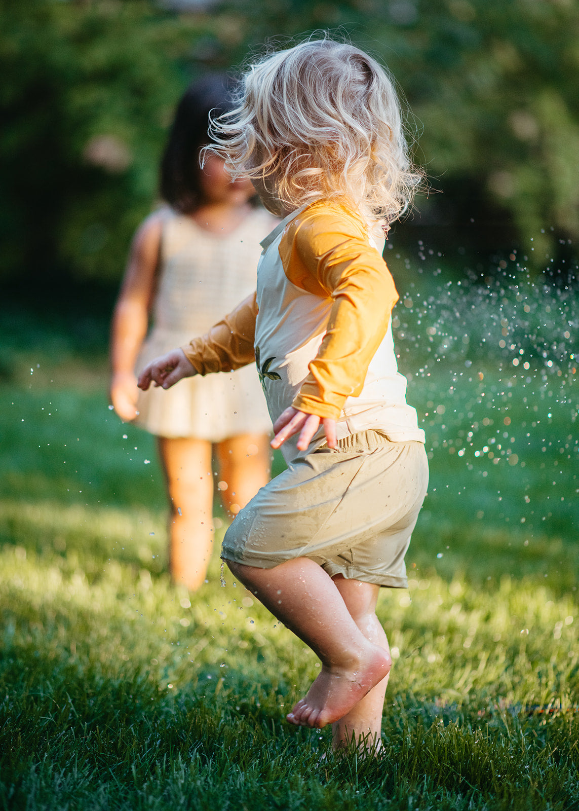 Boy running through the grass in Milkbarn Island Toucan long-sleeve shirt and trunks swim set, with girl in background wearing Milkbarn swimsuit, summer sprinkler play.