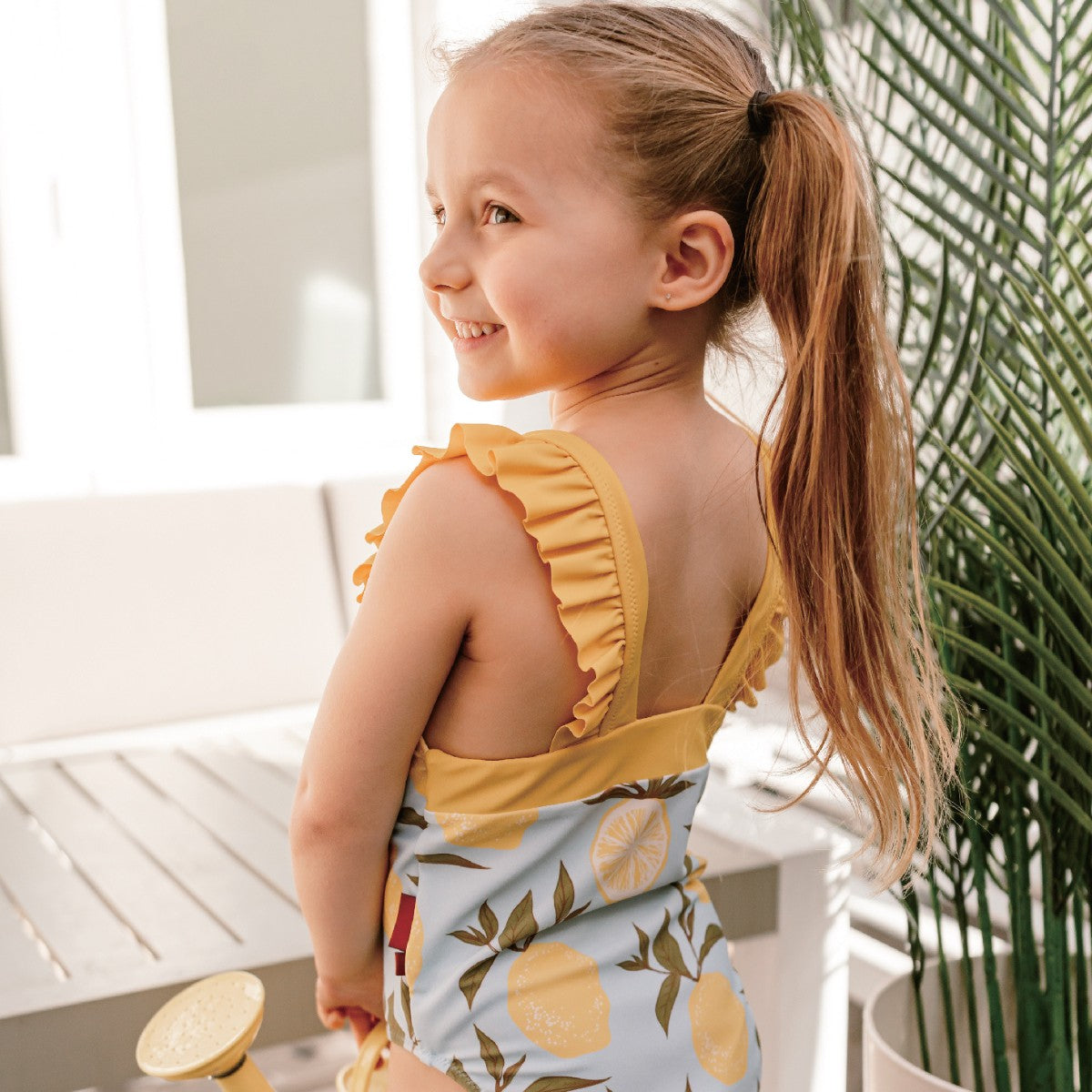 Girl wearing Milkbarn Lemon ruffle square-neck swimsuit with open back and ruffled straps, turning to smile at the camera, cute toddler swimwear.