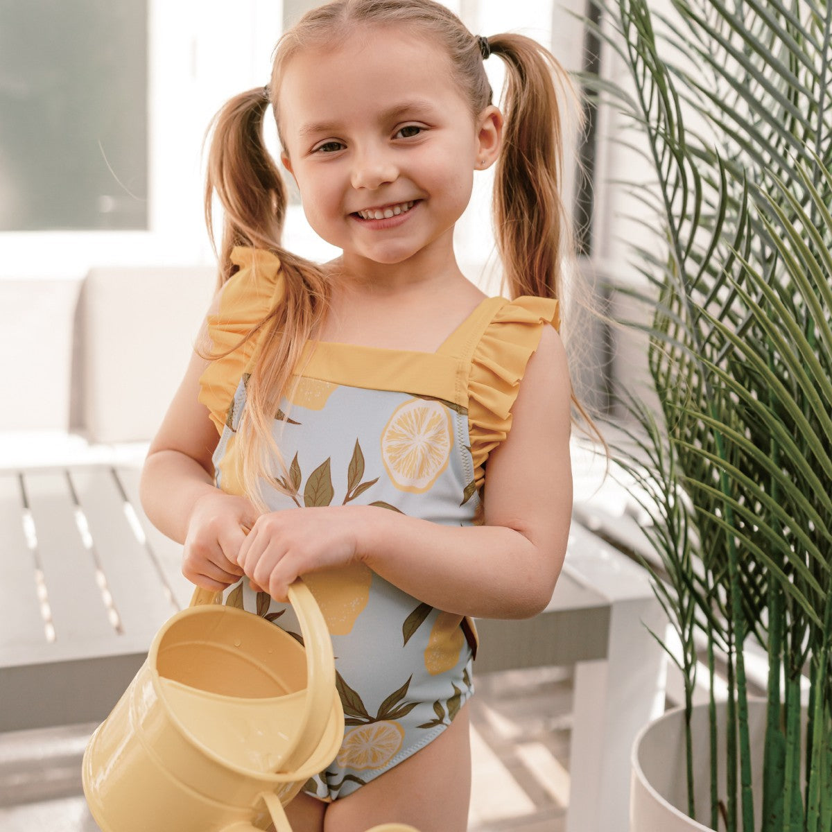 Close-up of young girl wearing Milkbarn Lemon ruffle square-neck swimsuit holding a yellow water pail, UPF 50+ baby swimwear for sunny play.