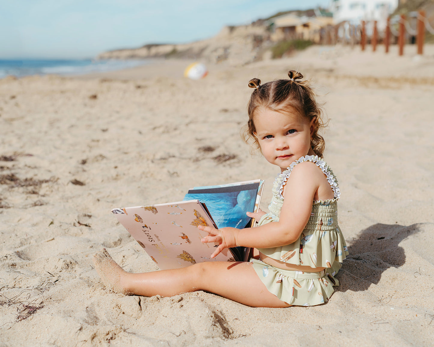 Girl sitting on the sand wearing Milkbarn Summer Surf ruffle two-piece swimsuit, reading A Sand Story, playful kids beachwear moment.