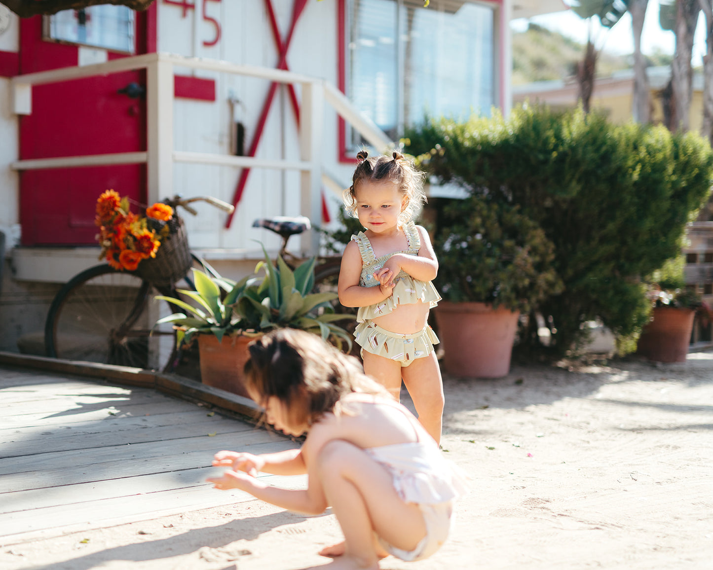 Two girls on a beach boardwalk wearing Milkbarn swimsuits, one in ruffle two-piece and one-piece, perfect for sunny summer adventures.