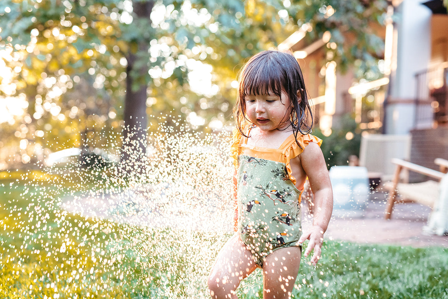 Girl playing in the grass under a water sprinkler wearing Milkbarn Toucan ruffle square-neck swimsuit, UPF 50+ kids swimwear with tropical bird print.