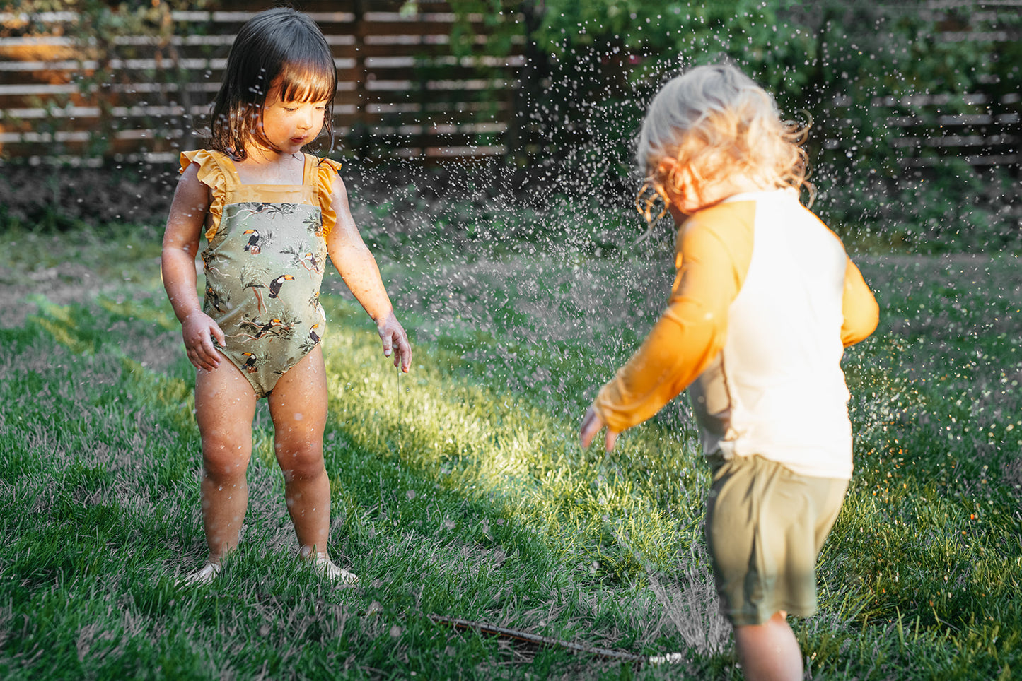 Girl and boy playing in the grass with backyard sprinkler, wearing Milkbarn Toucan swimsuits, ruffle square-neck for her and long-sleeve shirt and trunks for him.