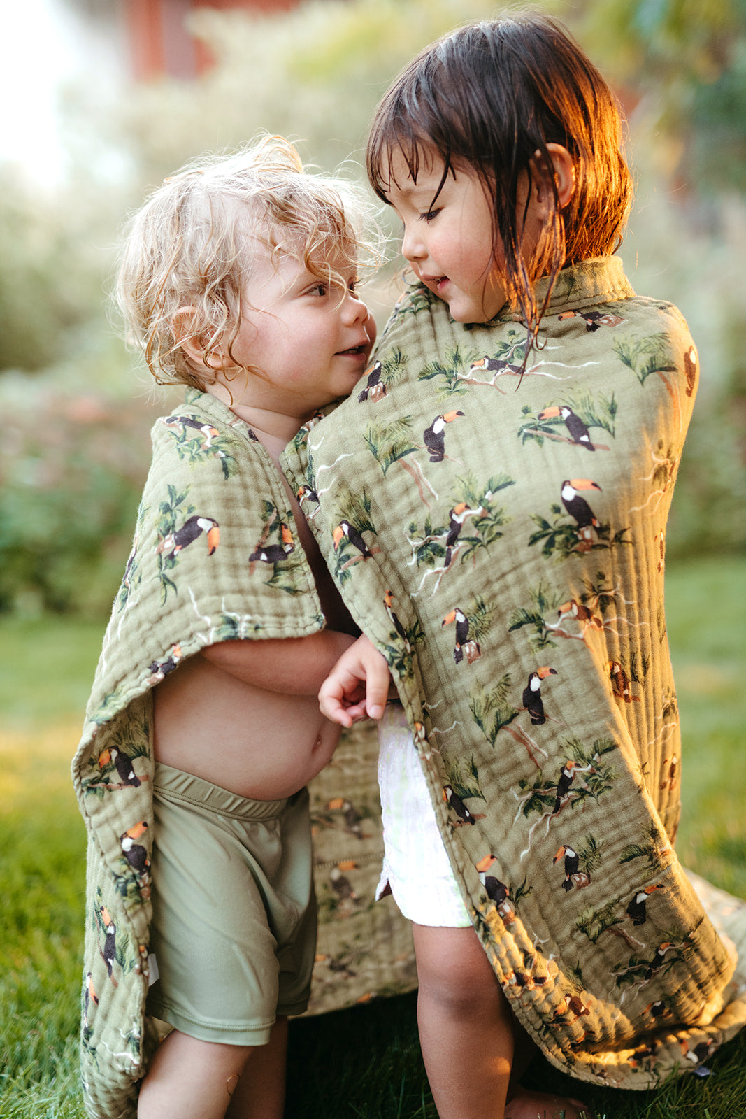 Young girl and boy standing side by side sharing Milkbarn Toucan organic cotton towel, playful summer moment after swimming or beach play.