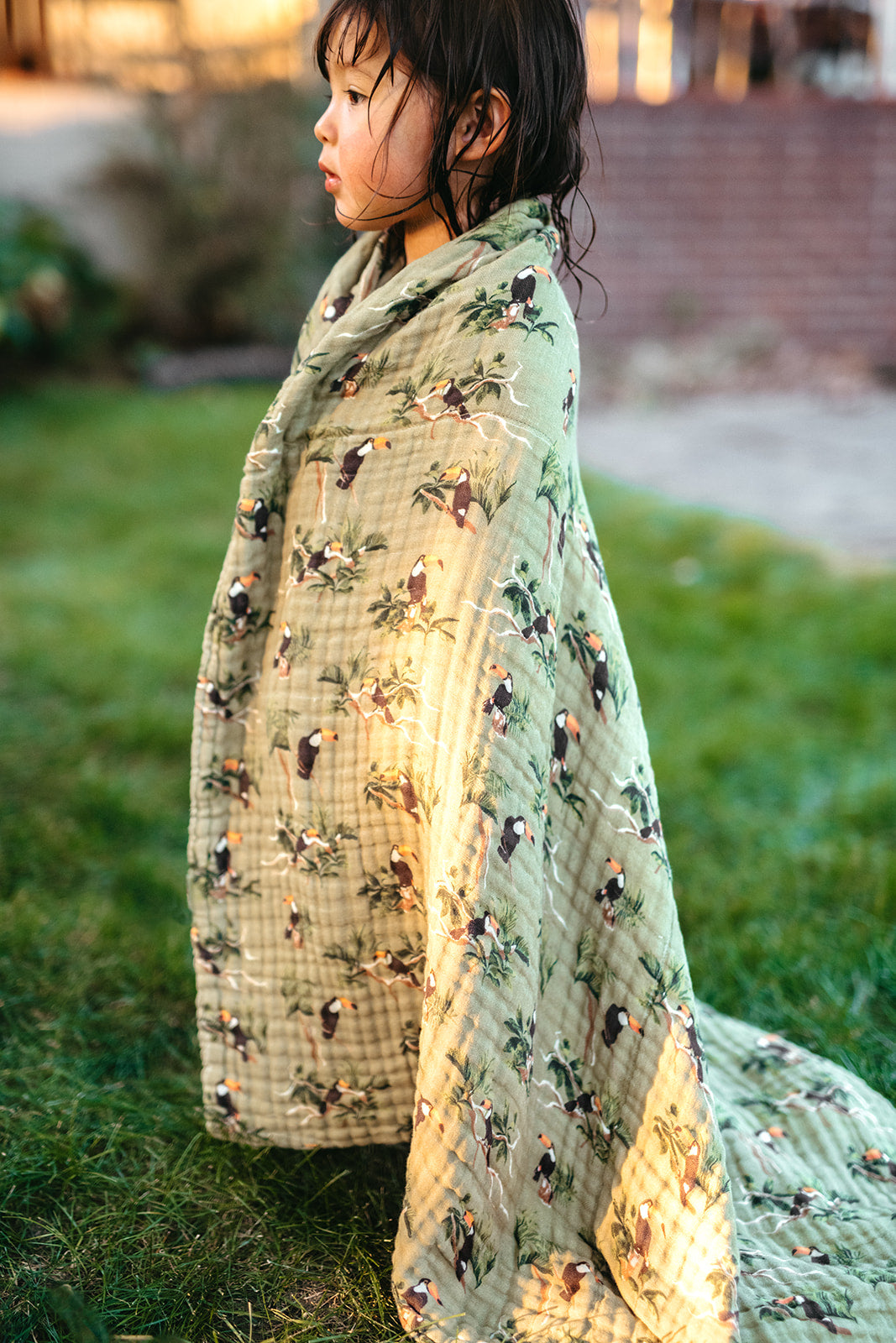 Little girl standing on the grass wrapped in Milkbarn Toucan cotton towel with tropical bird design, gentle and absorbent after bath or swim.