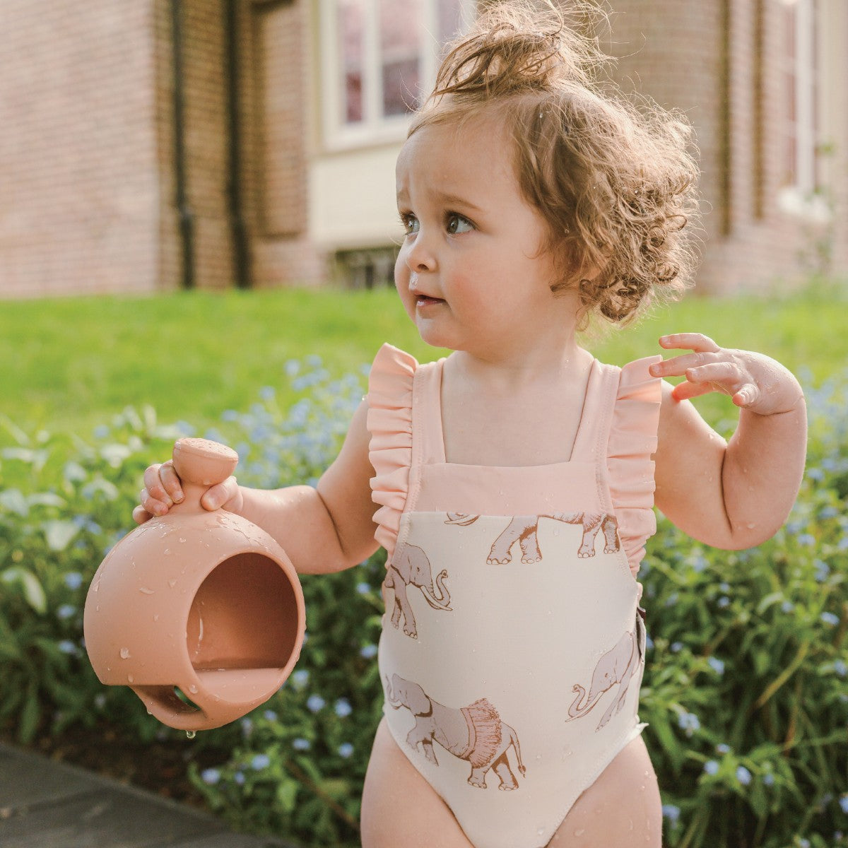 Little girl on the sidewalk wearing Milkbarn Tutu Elephant ruffle square-neck swimsuit holding a pink water toy, soft UPF 50+ baby swimwear.