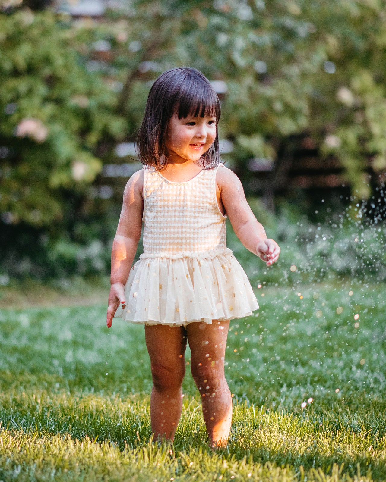 Little girl wearing Milkbarn’s Yellow Gingham Ballet Skirt Swimsuit splashes in backyard sprinklers, enjoying a bright summer day in a playful, vintage-inspired print