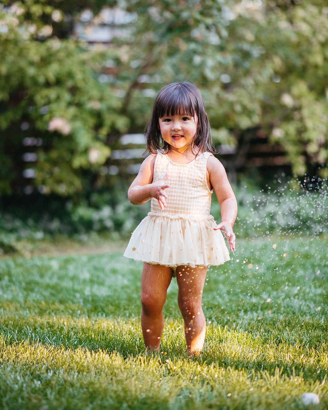 Smiling girl in Milkbarn’s Yellow Gingham Ballet Skirt Swimsuit twirls through backyard sprinklers, the soft yellow checks and tutu skirt capturing the joy of summer play