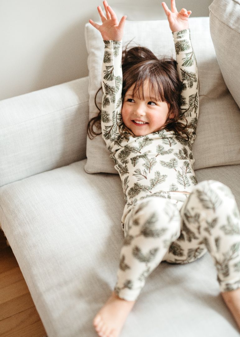 Little girl in Pine holiday bamboo cotton pajama set stretches on couch with hands in the air