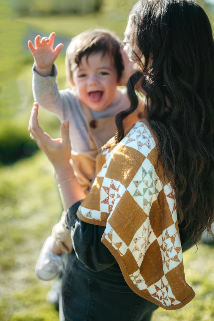 Baby boy smiling with hand raised while mom holds him in grassy field with Quilt print bamboo cotton burp cloth over shoulder