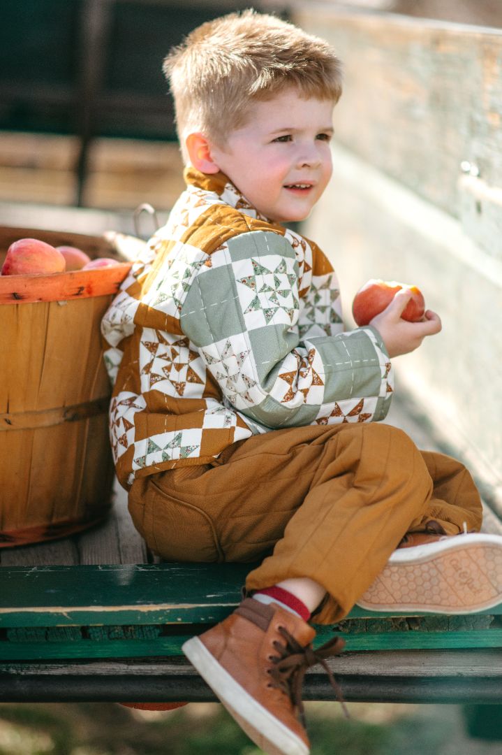 Side view of young boy in golden brown quilted pants sitting on wooden wagon in autumn setting