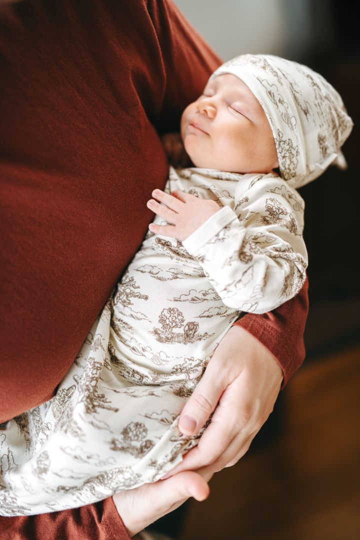 Sleeping baby held by mom, wearing a Country Toile print newborn gown and hat set in Luxe Stretch fabric made of 95% bamboo viscose and 5% spandex, featuring cows and farmland design.