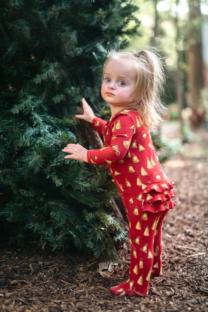 Baby girl in Vintage Bell ruffle footie stands beside pine tree in forest wearing holiday bamboo cotton pajamas
