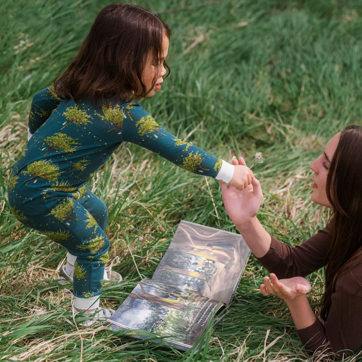 Baby girl and Mom playing in the grass reading the book Finn the Firefly by Wendy Wallace. The girl is wearing the Firefly Bamboo Zipper Pajama by Milkbarn