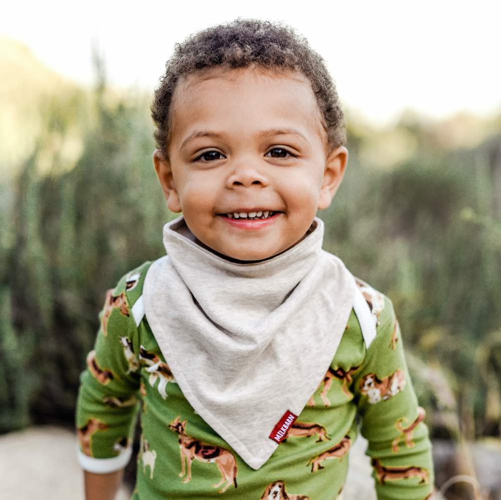 Little Boy on a Rock Wearing the Long Sleeve One Piece in the Green Dog Print and the Organic Kerchief Bib in the Heathered Oatmeal Fabric by Milkbarn Kids