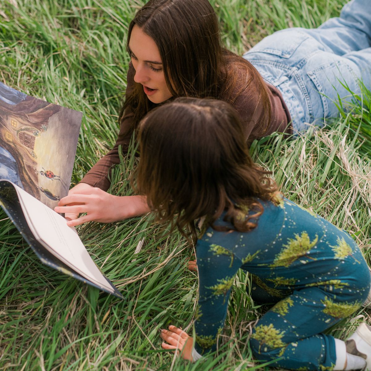 Mom and daughter sit on green grass read the Milkbarn children's book, Finn the Firefly by Wendy Wallace with illustrations by Haley Hunt. The baby girl wears the bamboo zipper PJs in the green Firefly print