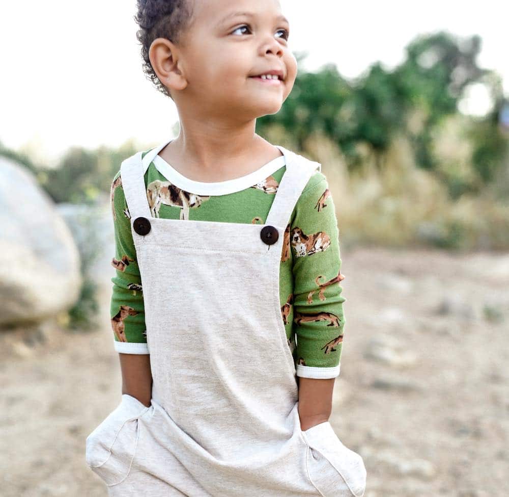 Little boy in the field wearing Milkbarn Kids organic Overall in Heathered Oatmeal fabric and a green dog Long Sleeve One Piece