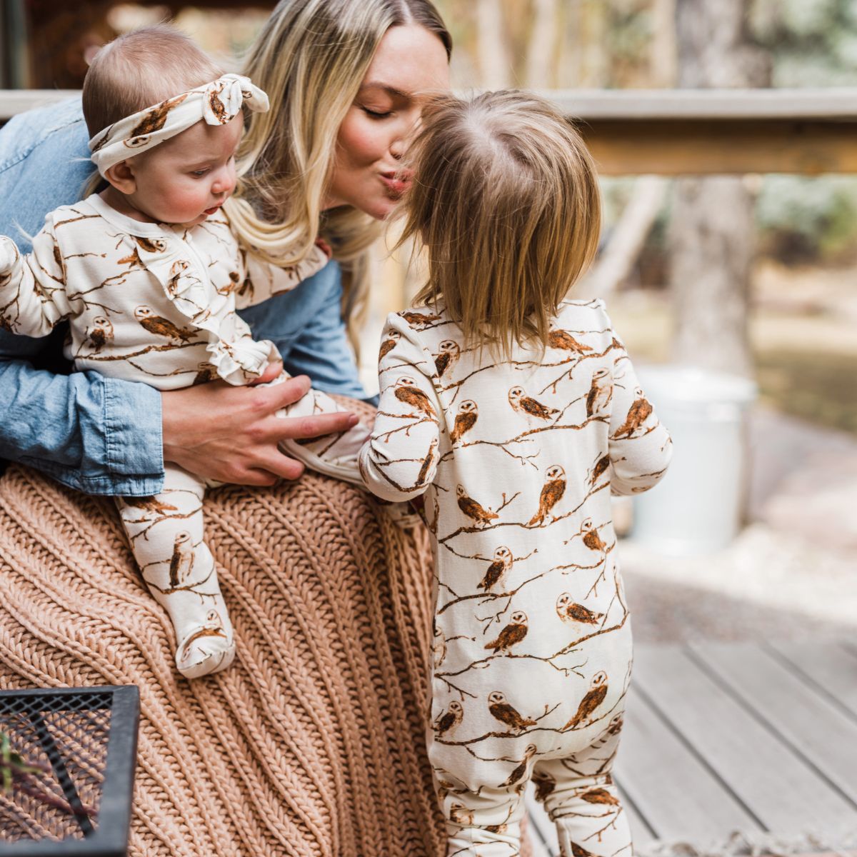 Mom holding a baby and leans over to kiss her son. He wears the owl zipper pajama by milkbarn kids