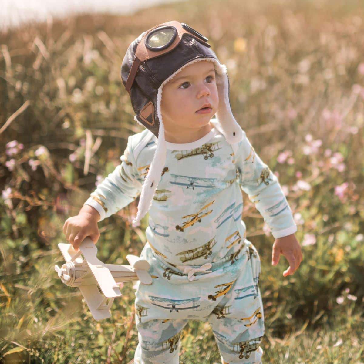 Baby boy looking into the sky while standing in a field near the ocean wearing an aviator hat and holding a toy vintage airplane while wearing the Vintage Planes Organic Long Sleeve One Piece and Jogger pants by Milkbarn