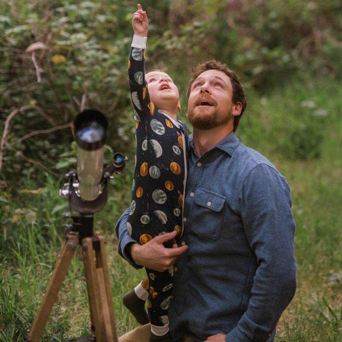 Baby boy and his Dad at dusk looking at the stars and planets with e telescope. The boy points up and wears the bamboo zipper pajamas in the planets print by Milkbarn Kids.