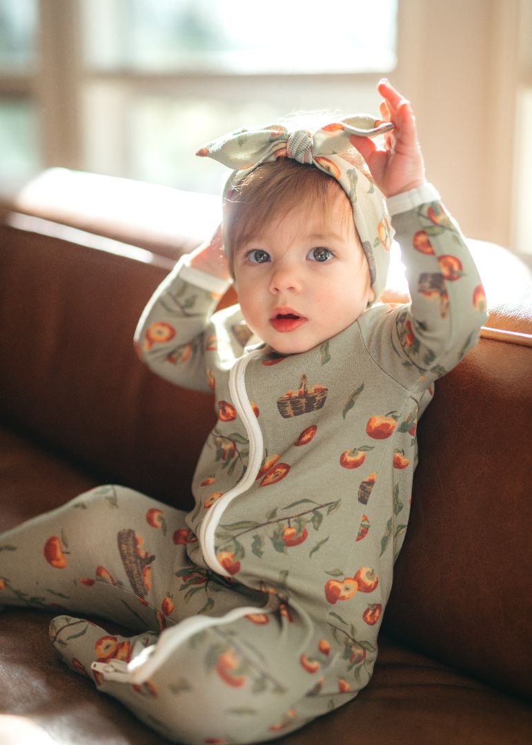 Toddler in Milkbarn Organic Cotton Apple Orchard print zipper footed romper and knotted headband, baby model sitting in cozy autumn baby clothes