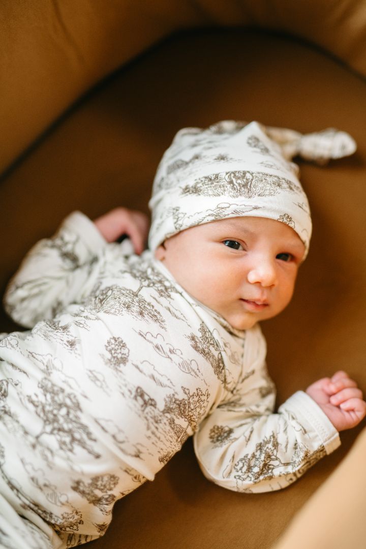 Awake baby lying in a crib, dressed in a Country Toile print newborn gown and hat set in breathable Luxe Stretch fabric made of 95% bamboo viscose and 5% spandex, with a cows and farmland pattern.
