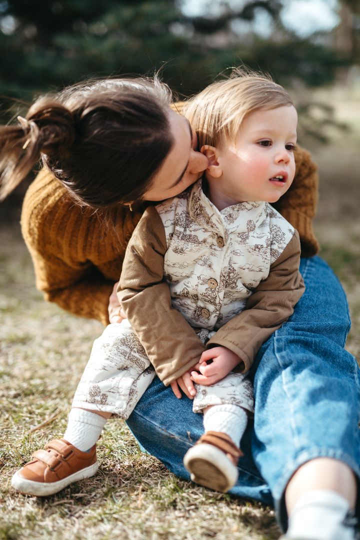 Little boy sitting and held by his mom, dressed in a warm quilted 100% cotton jumpsuit in our cow and farmland-themed Country Toile print, perfect for chilly weather.