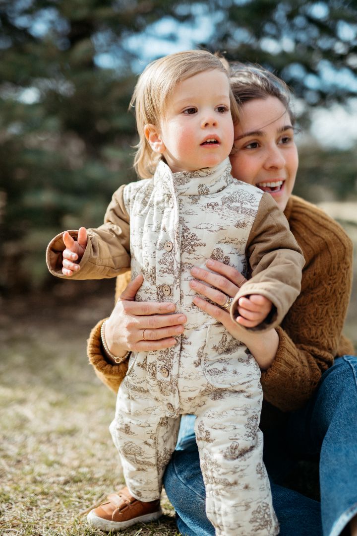 Little boy standing outside with his mom, wearing a 100% cotton quilted jumpsuit in our Country Toile print featuring cows and farmland, designed to keep warmth in during cold winter months.