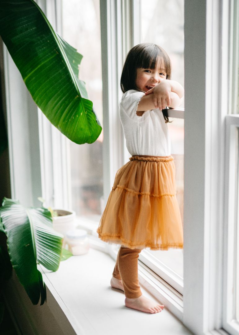 Girl facing the window in a gold tutu dress with attached leggings, crafted from a bamboo, cotton, and spandex blend for breathable comfort and playful elegance.