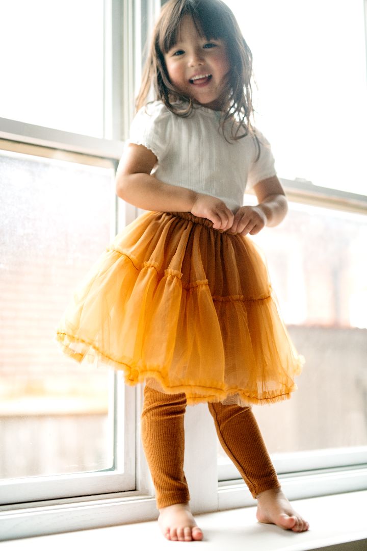 Girl standing in a window sill wearing a gold tulle tutu dress with attached leggings, made from a soft and stretchy blend of bamboo, cotton, and spandex.