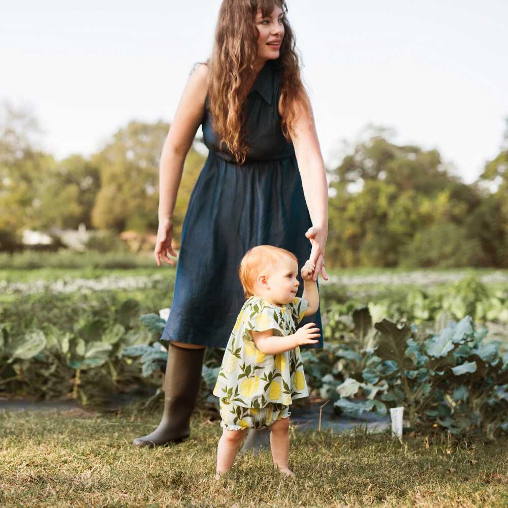 Lifestyle photo of baby girl wearing Milkbarn organic bamboo dress and bloomer outfit set in Lemon print, walking on a farm with her mom