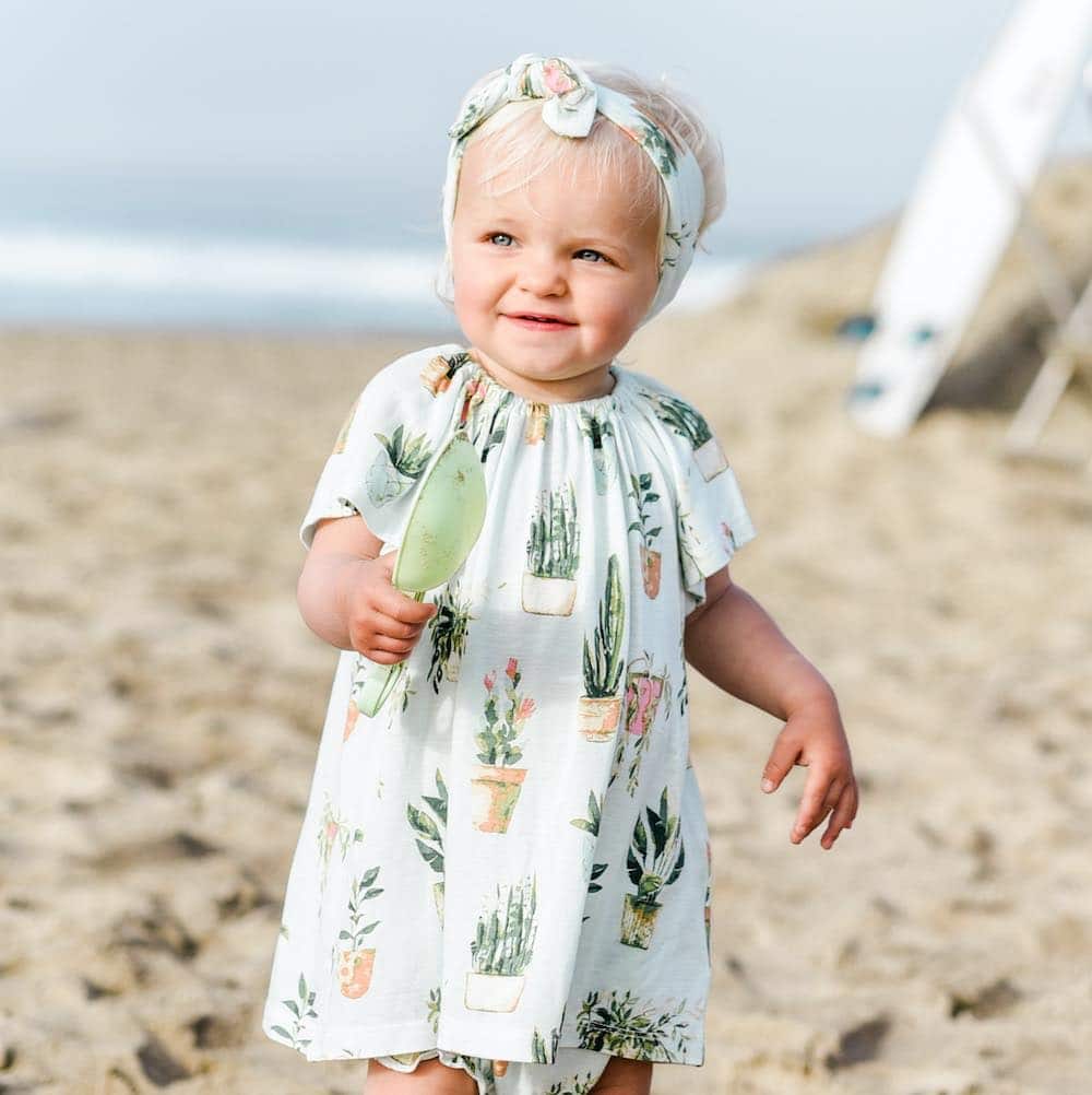 Lifestyle photo of toddler girl wearing Milkbarn organic bamboo dress and bloomer outfit set in Potted Plants print on the beach