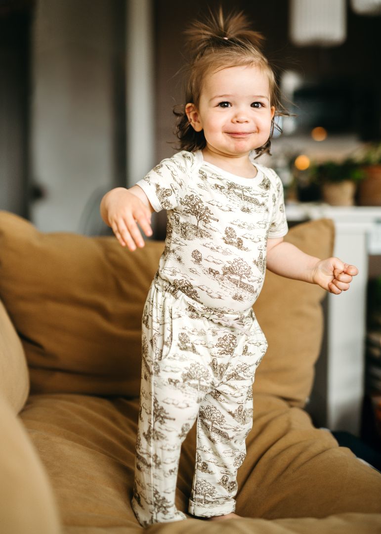 Child standing on a couch wearing jogger pants with an elastic waistband, drawstring, and pockets, paired with a Country Toile short sleeve one piece.