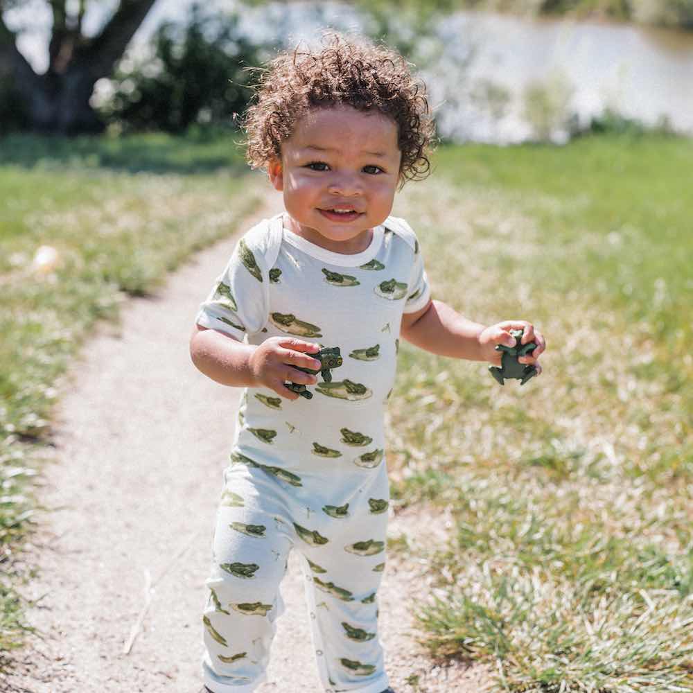 Little Baby in a Path in Front of a Lake Wearing a Bamboo Romper or Jumpsuit in the Leapfrog Print by Milkbarn Kids
