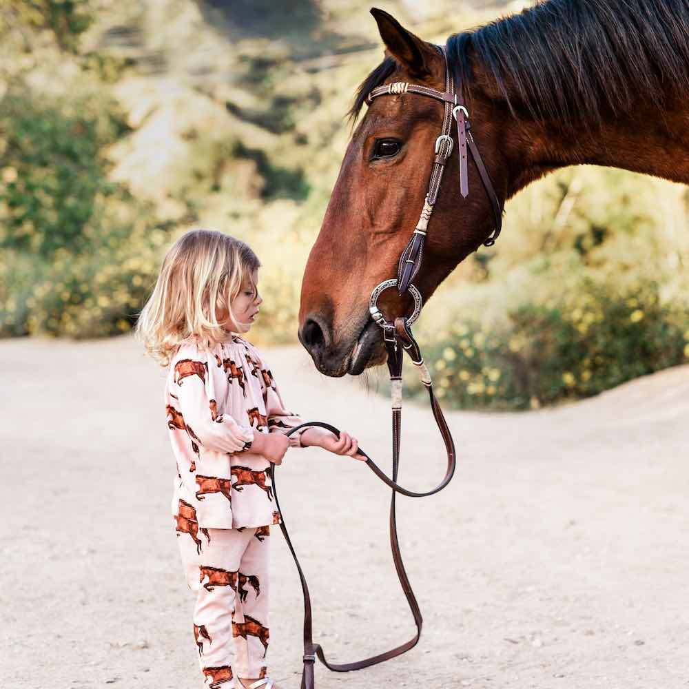 Little Girl Holding the Reins of a Horse and She Wears an Organic Cotton Long Sleeve Dress and Legging Set in the Horse Print by Milkbarn Kids