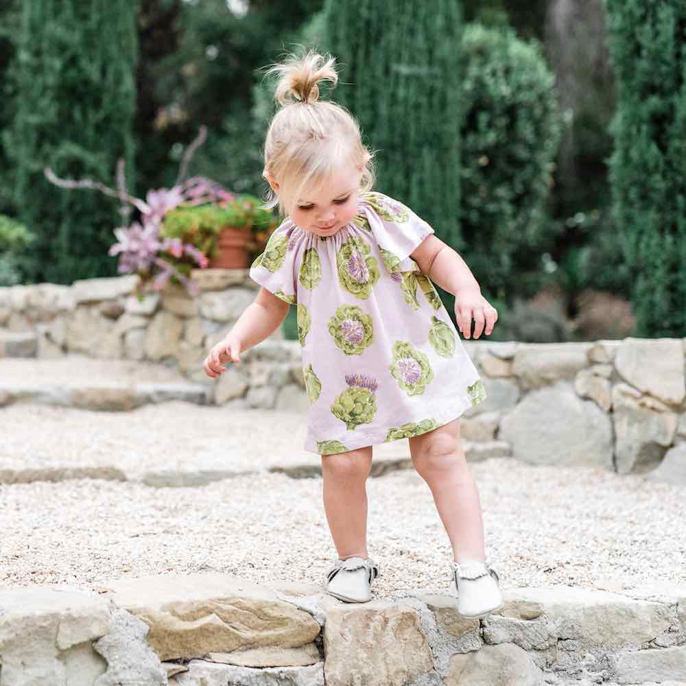 Little Girl Playing on Some Rocky Steps Wearing an Organic Cotton Dress and Bloomer Set in the Artichoke Vegetable Print by Milkbarn Kids