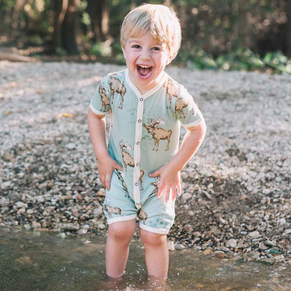 Baby boy in a river smiling wearing Milkbarn Kids Bamboo Shortall in the Blue Moose print