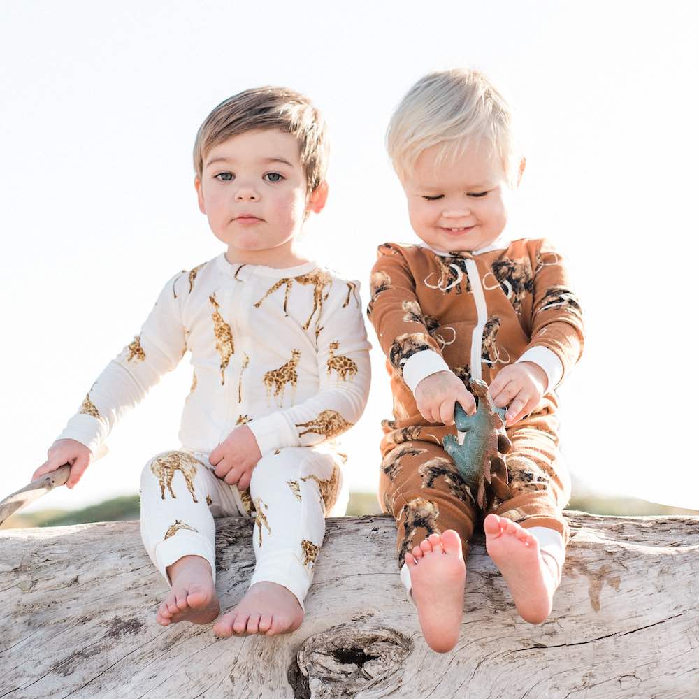 Two little baby boys wearing organic baby apparel by Milkbarn Kids consisting of a Zipper Pajama in Bamboo Orange Giraffe print and Organic Woolly Mammoth print