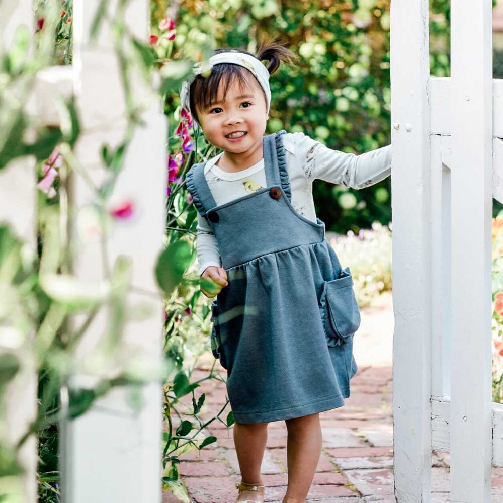 Little Baby Girl Opening a white Picket Fence Wearing a Dress Overall in the Organic and Recycled Denim by Milkbarn Kids