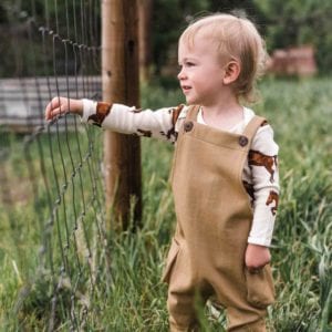 Little Baby Boy in a Field Wearing Organic Cotton and Rust Denim Overalls by Milkbarn Kids