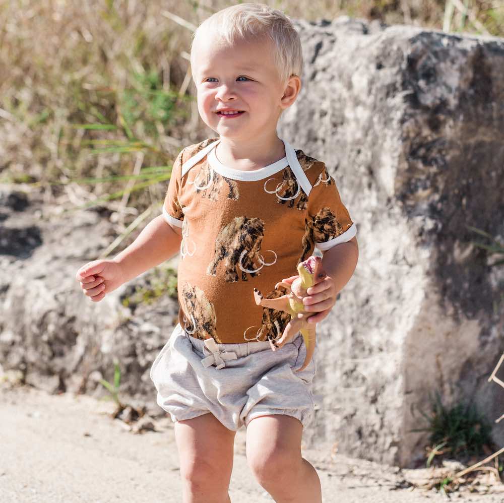Baby boy in a field wearing Milkbarn Kids organic Pocket Bloomer in Grey Pinstripe fabric and One Piece in Organic Woolly Mammoth Print