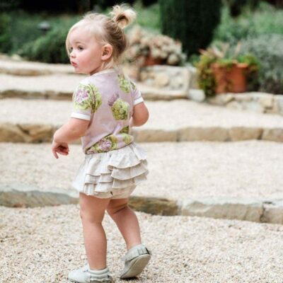 Baby Girl on Rock or Crushed Granite Steps Wearing the Ruffle Bloomer in the Organic Heathered Oatmeat and the Organic One Piece in the Artichoke Print by Milkbarn Kids