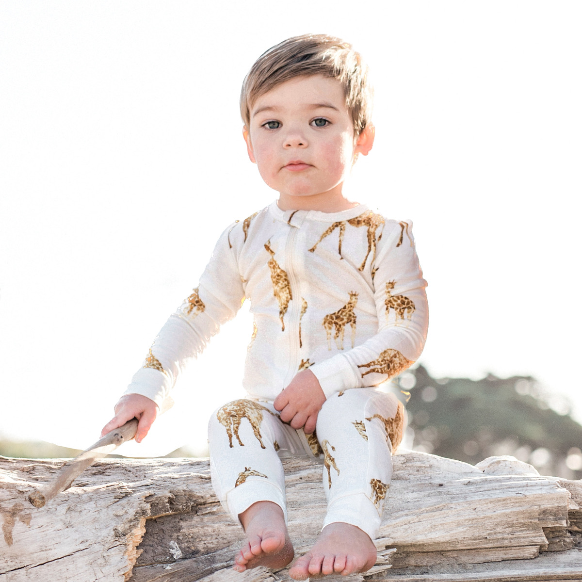 Baby boy sitting on a piece of drift wood log at the ocean holding a piece of drift while wearing the orange giraffe bamboo zipper pajamas by Milkbarn