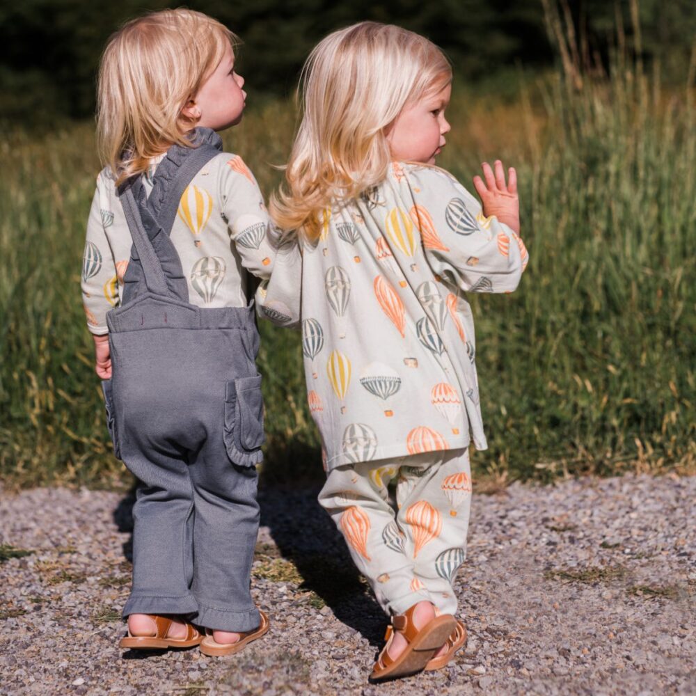Twins looknig out into a grassy field wearing the Denim Ruffle Overalls and the Vintage Balloons Long Sleeve Dress and Legging Set by Milkbarn
