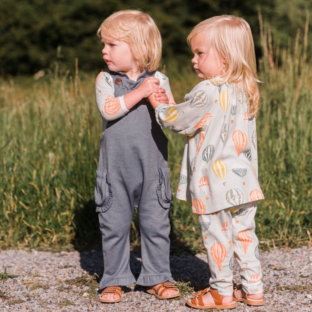 Twins holding hands in a grassy field while wearing the denim ruffle overalls and the Vintage Balloons Long Sleeve Dress and Legging set by Milkbarn
