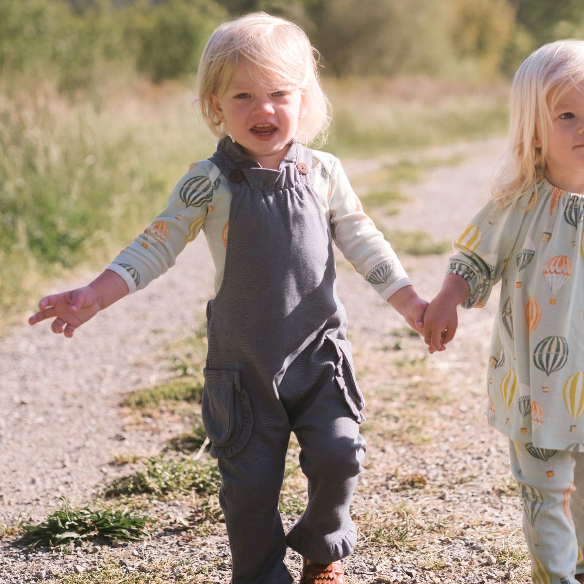 Twins walking on a dirt path wearing the Denim Ruffle Overalls by Milkbarn