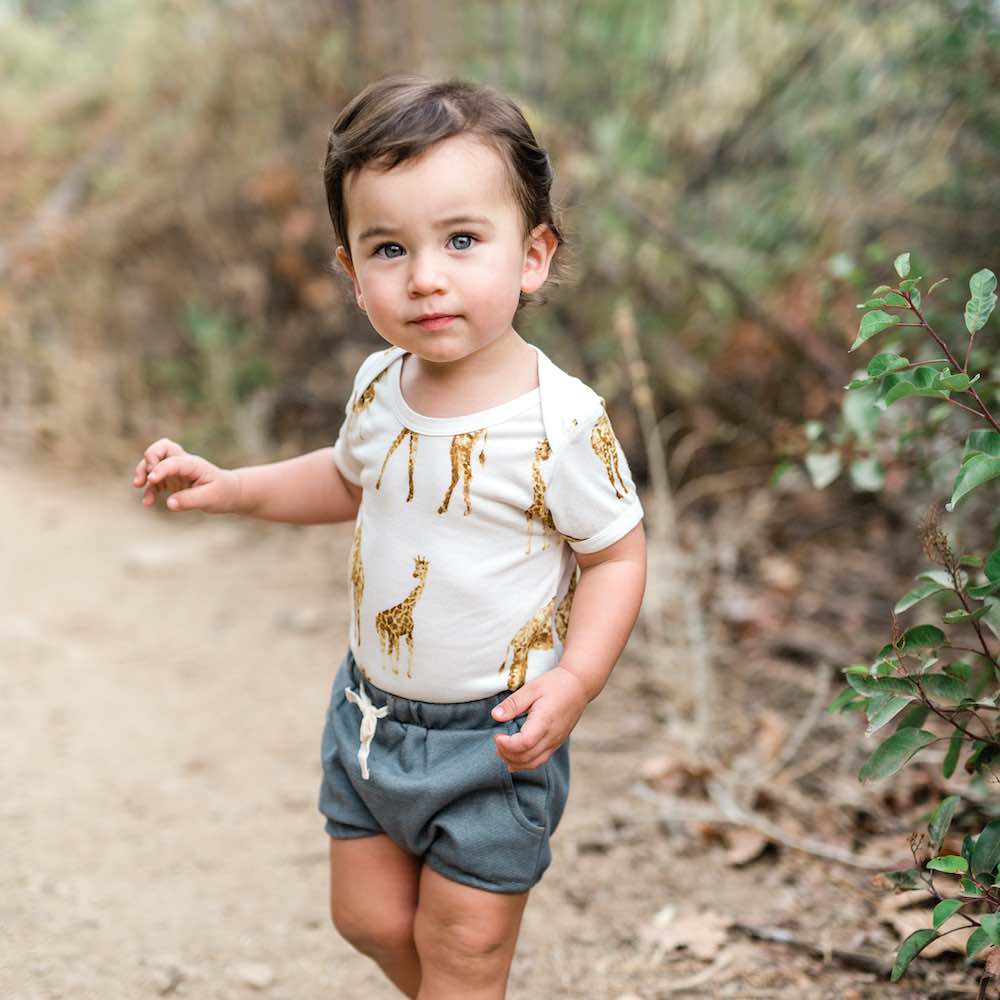 Baby Boy on a Dirt Path Wearing Denim Pocket Bloomers and the Bamboo One Piece in the Orange Giraffe Print by Milkbarn Kids